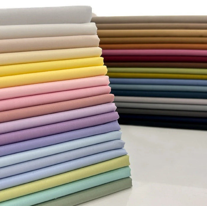 Des piles de Popeline polyester-coton au mètre de Longan Craft France, doux et respirant, dans des couleurs unies allant des pastels aux tons de terre, disposées sur une surface réfléchissante. Idéal pour robes, chemises et nappes.