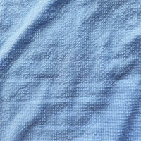 Un gros plan du polyester Seersucker à carreaux vichy au mètre de Longan Craft France montre sa surface texturée et froissée bleu clair, avec un motif gaufré subtil et de légers plis sur le tissu.