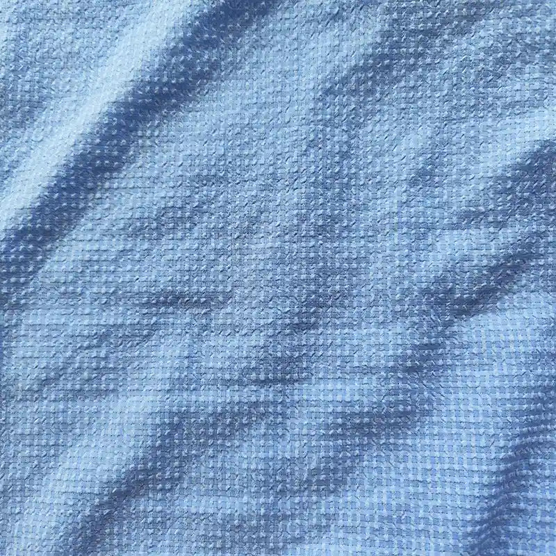 Un gros plan du polyester Seersucker à carreaux vichy au mètre de Longan Craft France montre sa surface texturée et froissée bleu clair, avec un motif gaufré subtil et de légers plis sur le tissu.