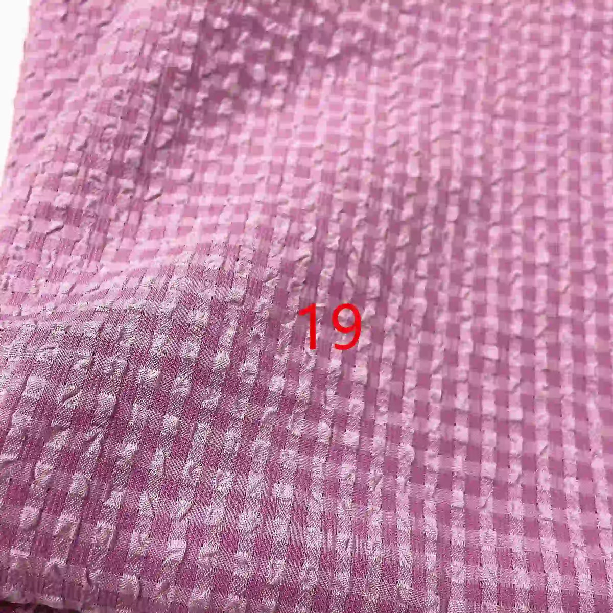 Le Seersucker polyester à carreaux vichy au mètre de Longan Craft France est un tissu rose texturé et froissé avec un motif en grille. Le chiffre "19" apparaît en rouge au centre de l'image.