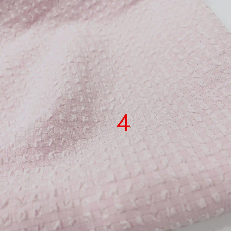 Gros plan sur le polyester Seersucker à carreaux vichy au mètre de Longan Craft France, montrant un tissu rose pâle froissé avec un motif carré tissé et un numéro 4 rouge imprimé près du centre.