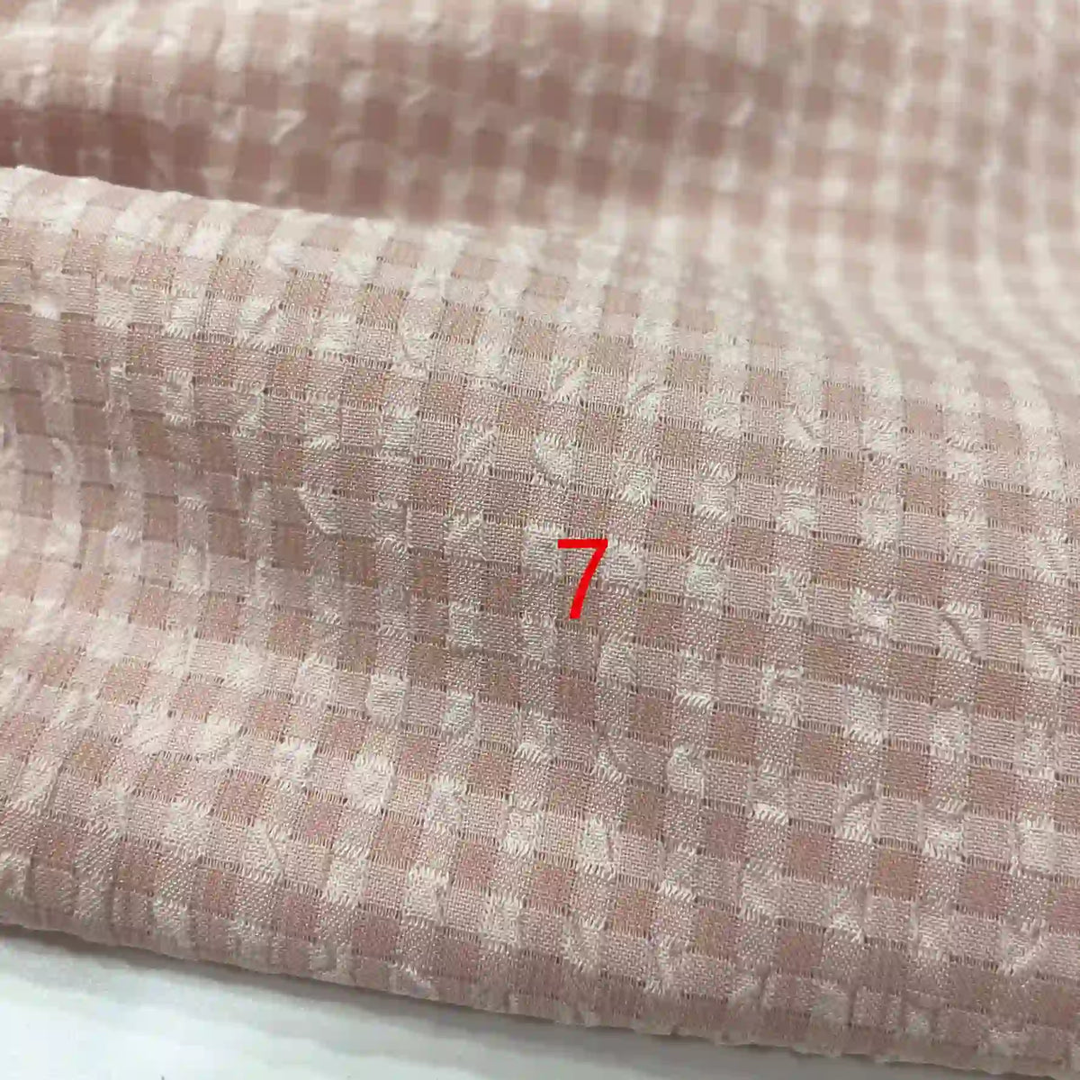 Un gros plan du polyester seersucker à carreaux vichy de Longan Craft France montre un tissu texturé rose clair et blanc avec un numéro 7 rouge centré.