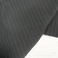 Un gros plan de Seersucker à larges rayures au mètre de Longan Craft France montre un tissu de polyester seersucker gris foncé subtilement rayé pour chemises, posé à plat sur du blanc avec des bords visibles et une petite section effilochée.