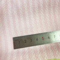 Une règle en métal argenté (1-6 cm, 5 en rouge) est posée sur le Seersucker à larges rayures au mètre de Longan Craft France, un tissu seersucker vintage en polyester arc-en-ciel pour chemises. Le tissu rose pâle présente des rayures horizontales respirantes.