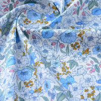 Un gros plan du tissu popeline de coton imprimé au mètre de Longan Craft France montre des motifs floraux bleus, jaunes et roses avec des feuilles vertes sur fond blanc. Ce tissu 100% coton, doux et légèrement froissé, est idéal pour le bricolage, le quilting, les langes, les peignoirs et les chemises.