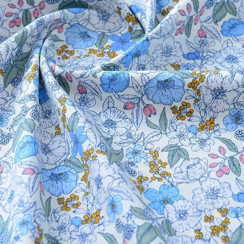 Un gros plan du tissu popeline de coton imprimé au mètre de Longan Craft France montre des motifs floraux bleus, jaunes et roses avec des feuilles vertes sur fond blanc. Ce tissu 100% coton, doux et légèrement froissé, est idéal pour le bricolage, le quilting, les langes, les peignoirs et les chemises.