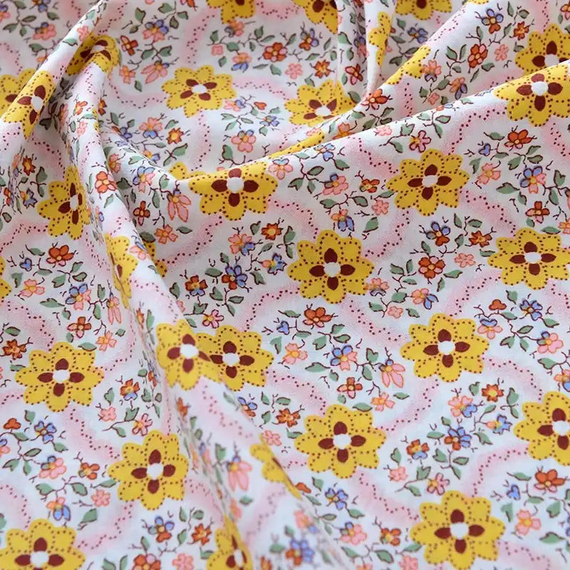 Un gros plan de Tissu popeline de coton imprimé au mètre par Longan Craft France montre des motifs floraux jaunes, orange et roses avec des feuilles vertes sur un fond clair ; le tissu 100 % coton est légèrement froissé et légèrement drapé.