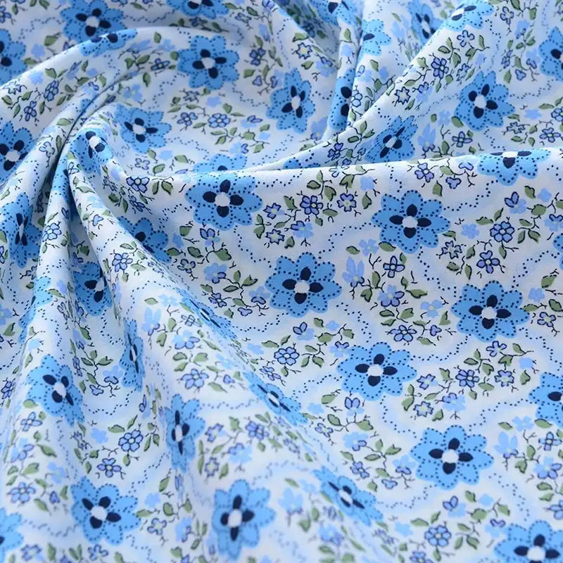 Gros plan sur le tissu popeline de coton imprimé au mètre de Longan Craft France, 100% coton à motifs floraux bleus et verts sur fond bleu clair, légèrement froissé et idéal pour DIY, quilting, nappes, robes ou chemises.