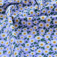 Un gros plan du Tissu popeline de coton imprimé au mètre de Longan Craft France montre des marguerites blanches et jaunes avec des feuilles vertes sur un tissu 100% coton violet clair, parfait pour le bricolage, le quilting, les couches, les peignoirs et les chemises.