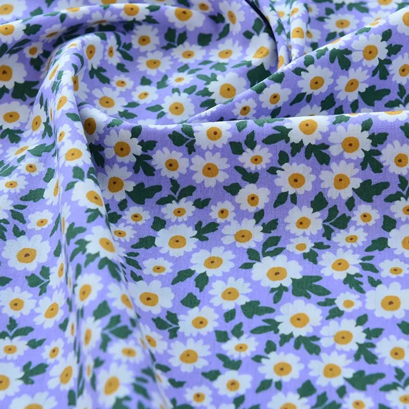 Un gros plan du Tissu popeline de coton imprimé au mètre de Longan Craft France montre des marguerites blanches et jaunes avec des feuilles vertes sur un tissu 100% coton violet clair, parfait pour le bricolage, le quilting, les couches, les peignoirs et les chemises.