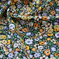 Gros plan sur le Tissu popeline de coton imprimé au mètre de Longan Craft France, qui présente un motif floral vibrant - fleurs jaunes, blanches, orange et violet clair sur fond noir - idéal pour le bricolage, le quilting, les langes, les peignoirs et les chemises. Légèrement froissé pour plus de texture.