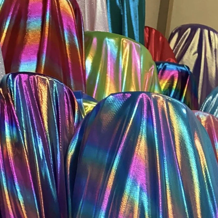 Les objets drapés dans le Tissu lamé arc-en-ciel au mètre de Longan Craft France - un tissu laser en polyester lamé rayé irisé - scintillent en bleu, violet, vert et rose, reflétant la lumière avec un effet arc-en-ciel éblouissant et masquant les costumes de scène qui se trouvent en dessous.