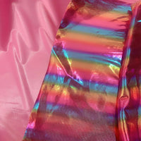 Une bande de Tissu lamé arc-en-ciel au mètre de Longan Craft France, polyester irisé à rayures dégradées, reflète des teintes métalliques vives à côté d'un tissu rose mat-parfait pour costumes de scène ou événements spéciaux.