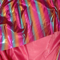 Gros plan sur deux tissus : la couche supérieure est du Tissu lamé arc-en-ciel au mètre de Longan Craft France, un polyester irisé à rayures dégradées idéal pour les costumes de scène, sur un tissu rose mat.