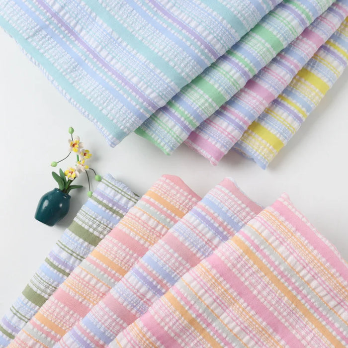 Six pièces de tissu seersucker rayé arc-en-ciel de Longan Craft France sont disposées en éventail sur une surface blanche, un petit vase sombre contenant des fleurs artificielles blanches et jaunes étant placé à gauche des tissus.