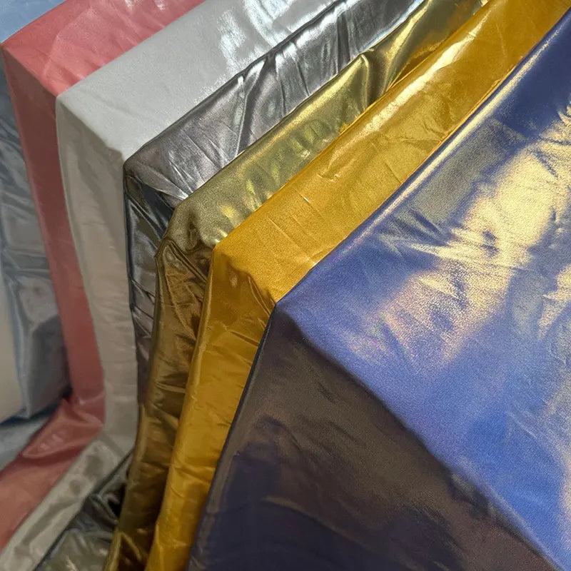 Plusieurs feuilles de Tissu satin lamé au mètre de Longan Craft France en rose, argent, or, jaune et bleu montrent leur éclat métallique - idéal pour les robes DIY, les costumes de scène ou les projets créatifs de couture.