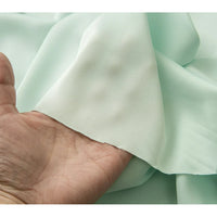 Une main soulève un morceau de Tissu mousseline satinée au mètre de Longan Craft France, montrant sa texture lisse, vert pastel, légèrement transparente - une mousseline soyeuse extensible et douce, idéale pour créer des robes de soirée élégantes.