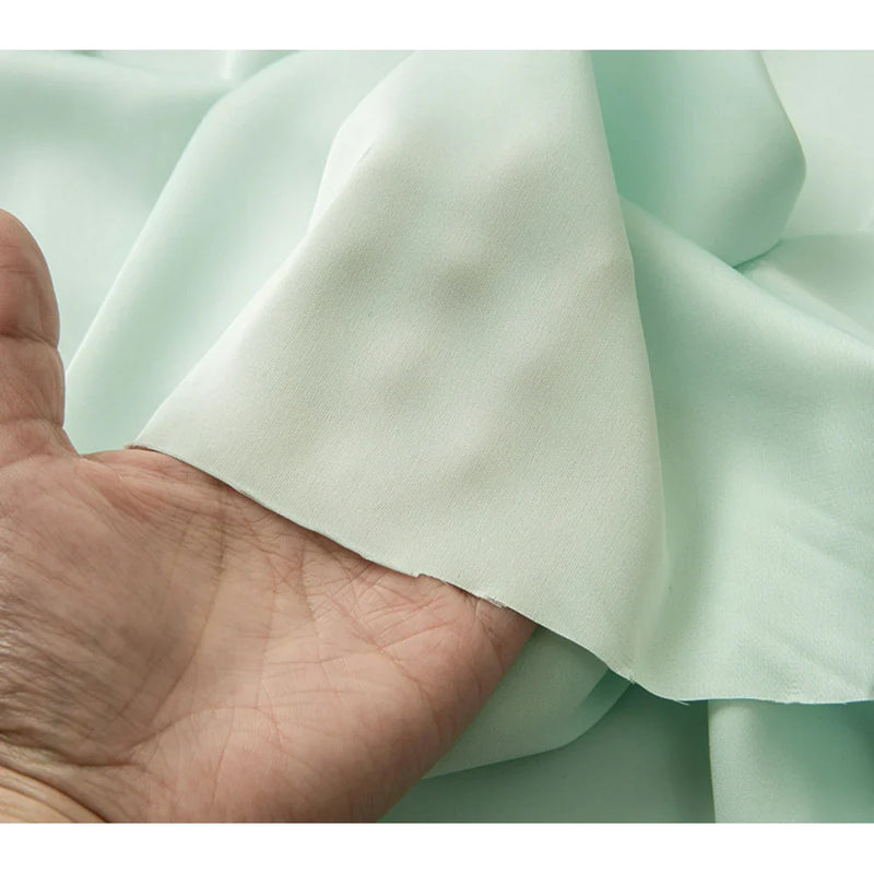 Une main soulève un morceau de Tissu mousseline satinée au mètre de Longan Craft France, montrant sa texture lisse, vert pastel, légèrement transparente - une mousseline soyeuse extensible et douce, idéale pour créer des robes de soirée élégantes.
