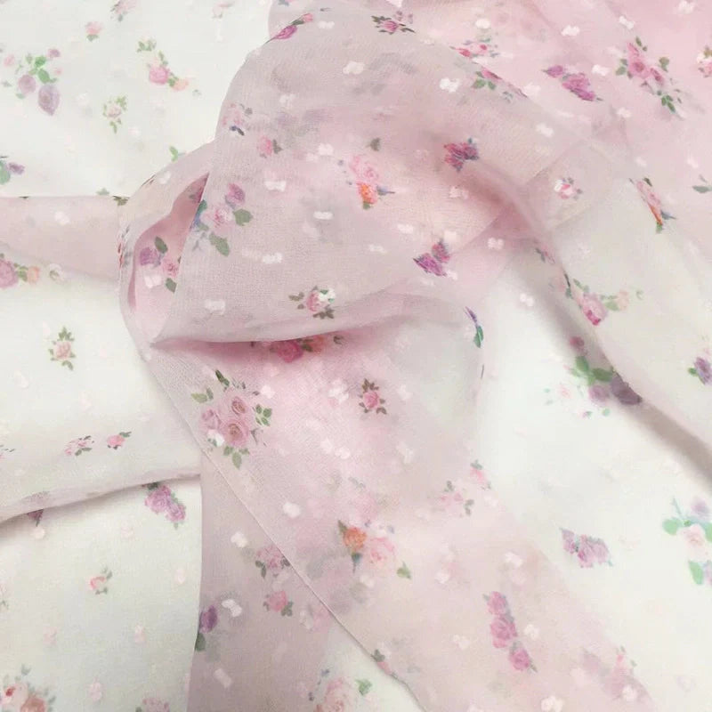 Un gros plan du tissu mousseline à petites fleurs au mètre de Longan Craft France montre son délicat imprimé floral rose, violet et vert sur un tissu diaphane aéré, parfait pour les robes d'été élégantes et les projets créatifs.