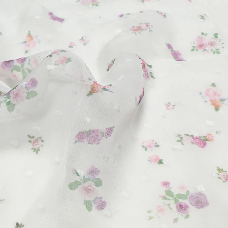 Le tissu mousseline à petites fleurs au mètre de Longan Craft France : des plumetis blancs transparents à la texture subtilement pointillée, imprimés de petites roses roses et violettes et de feuilles vertes - parfaits pour les robes d'été.
