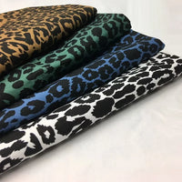 Quatre Tissu jean imprimé léopard 10oz de Longan Craft France - 100 % coton denim - sont empilés en rangée, chacun avec un motif de couleur différente : brun/noir, vert/noir, bleu/noir et blanc/noir, sur un fond uni.