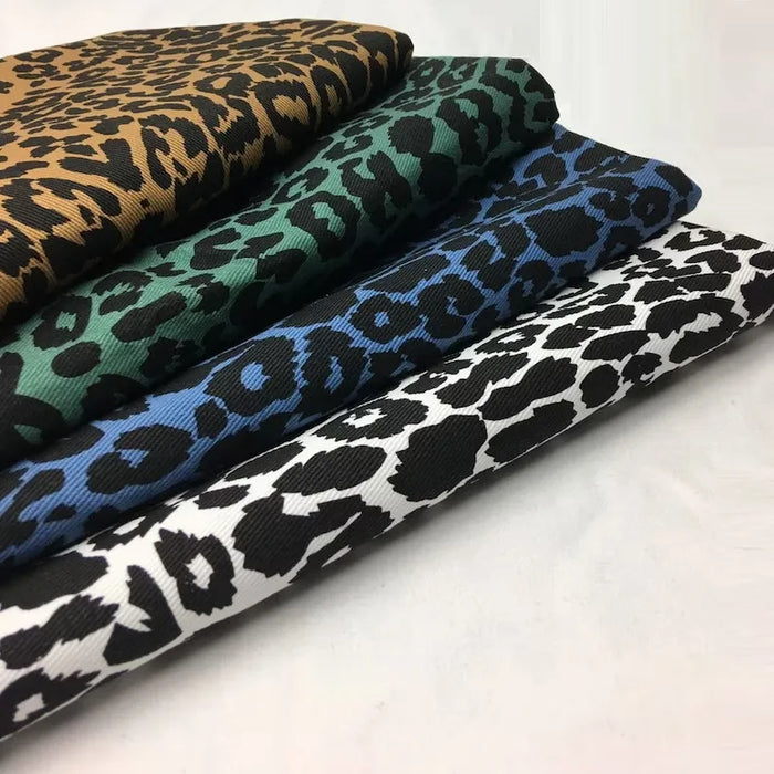 Quatre Tissu jean imprimé léopard 10oz de Longan Craft France - 100 % coton denim - sont empilés en rangée, chacun avec un motif de couleur différente : brun/noir, vert/noir, bleu/noir et blanc/noir, sur un fond uni.