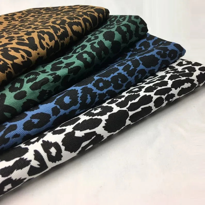 Quatre Tissu jean imprimé léopard 10oz de Longan Craft France - 100 % coton denim - sont empilés en rangée, chacun avec un motif de couleur différente : brun/noir, vert/noir, bleu/noir et blanc/noir, sur un fond uni.