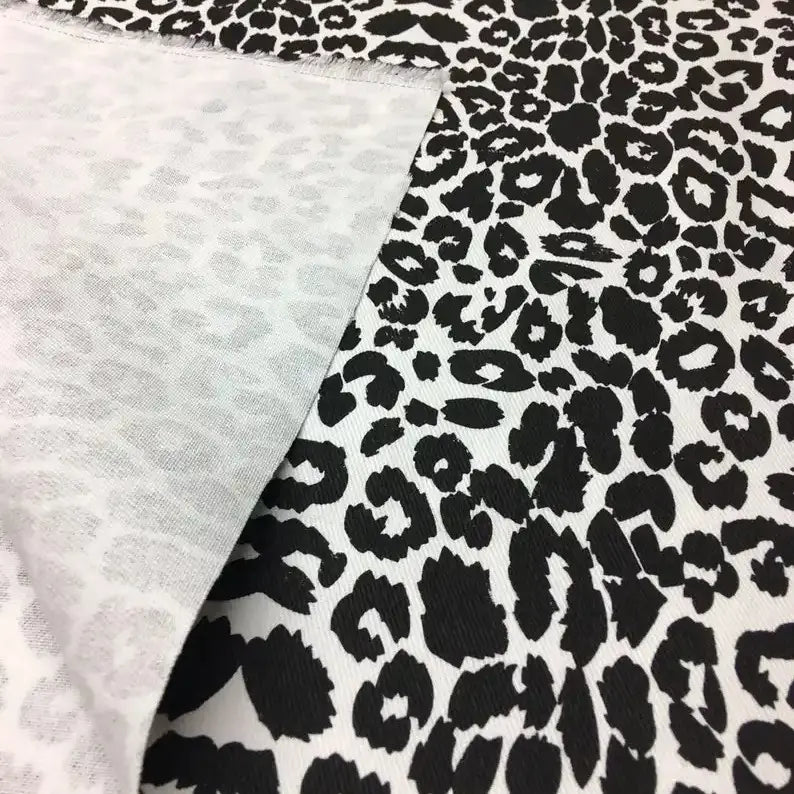 Gros plan sur le Tissu jean imprimé léopard 10oz de Longan Craft France, un denim 100 % coton délavé avec un imprimé léopard noir et blanc, dont un coin est retourné pour révéler le motif inversé, plus clair et moins distinct.
