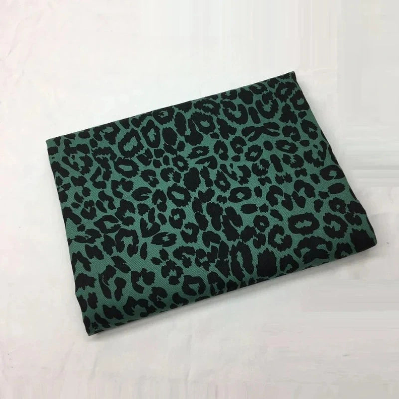 Un morceau de Tissu jean imprimé léopard 10oz de Longan Craft France, présentant un fond vert avec un motif léopard noir, est présenté sur une surface blanche unie.