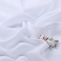 Une petite figurine de chat en céramique avec des marques rouges et noires repose sur la mousseline légère transparente au mètre de Longan Craft France, un tissu polyester fin et doux qui ajoute une délicate élégance à la scène.
