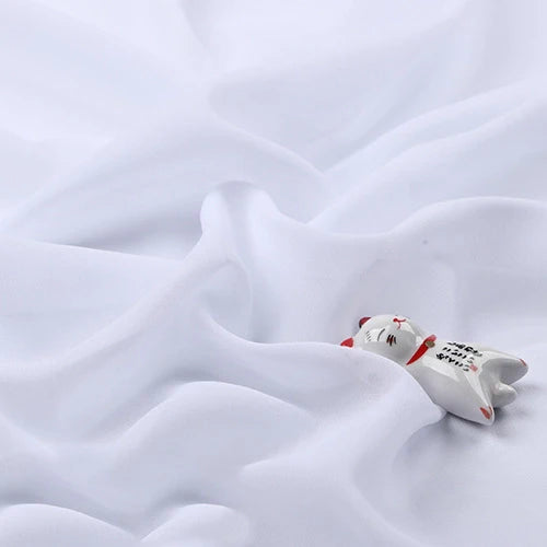Une petite figurine de chat en céramique avec des marques rouges et noires repose sur la mousseline légère transparente au mètre de Longan Craft France, un tissu polyester fin et doux qui ajoute une délicate élégance à la scène.