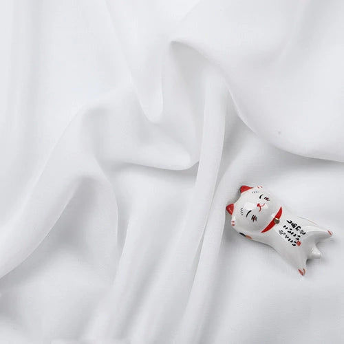 Un petit chat blanc en céramique avec des oreilles rouges et des marques noires repose sur la Mousseline légère transparente au mètre de Longan Craft France, une mousseline en tissu de polyester fin et doux, créant un arrière-plan élégant et délicat.