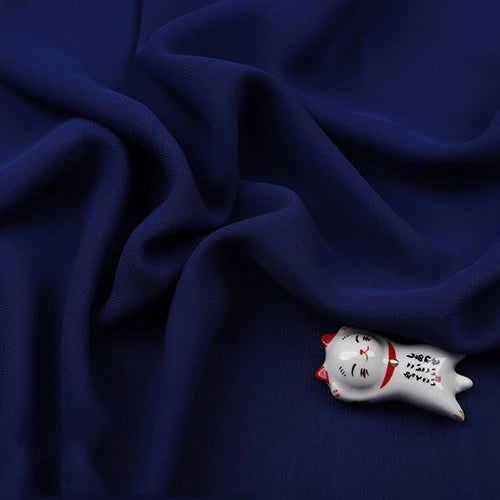 Un petit chat en céramique aux oreilles rouges et à l'écriture japonaise repose sur un tissu polyester doux et transparent "Mousseline légère" de Longan Craft France. La mousseline bleu foncé est légèrement froissée, mettant en valeur la figurine.