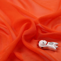 Mousseline légère transparente au mètre" de Longan Craft France, orange vif, drapée de plis souples, à côté d'une petite figurine de chat en céramique blanche avec des détails rouges et noirs.