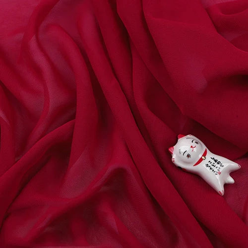 Une petite figurine de chat blanc avec des marques rouges et une écriture noire repose sur de la Mousseline légère transparente au mètre de Longan Craft France, créant un effet délicat et semi-transparent.
