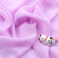 Une petite figurine de chat en céramique aux traits peints repose sur le doux et fin tissu mousseline polyester au mètre de Longan Craft France, une mousseline légère et transparente, drapée d'un doux rose pâle.