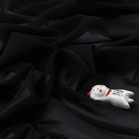 Un petit chat blanc en céramique avec des accents rouges repose sur une "Mousseline légère transparente au mètre" de Longan Craft France - un tissu en polyester fin et doux avec une finition mate et des plis élégants.