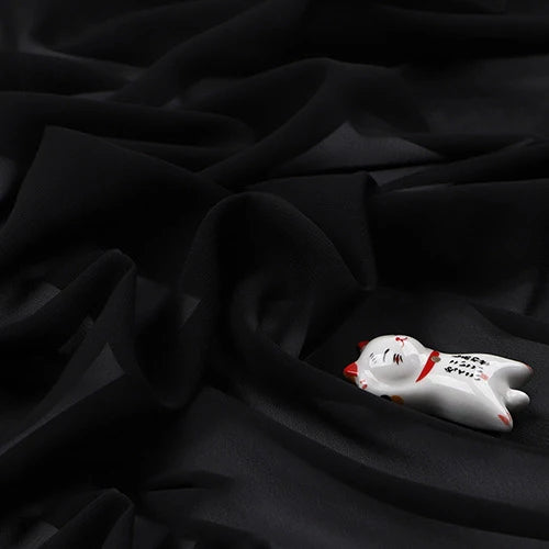 Un petit chat blanc en céramique avec des accents rouges repose sur une "Mousseline légère transparente au mètre" de Longan Craft France - un tissu en polyester fin et doux avec une finition mate et des plis élégants.