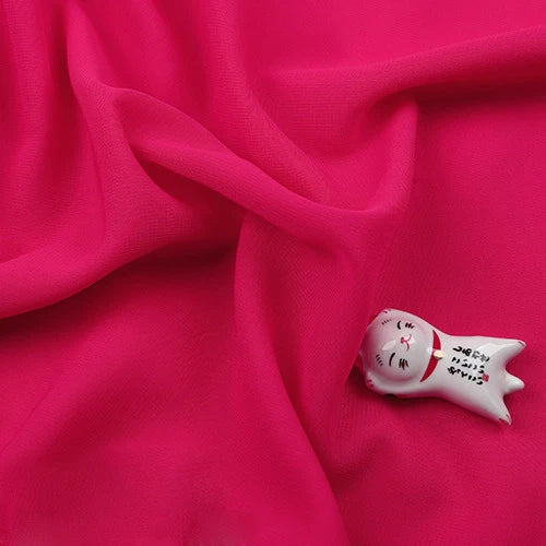 Une "Mousseline légère transparente au mètre" de Longan Craft France, d'un rose vif et doux, est délicatement drapée. Dans le coin inférieur droit, une petite figurine de chat en céramique blanche avec un sourire peint repose sur le tissu délicat.