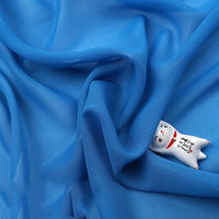Une Mousseline légère bleue transparente au mètre de Longan Craft France est drapée en plis souples, recouvrant partiellement une petite figurine de chat en céramique blanche peinte avec des traits de visage et une écriture japonaise.