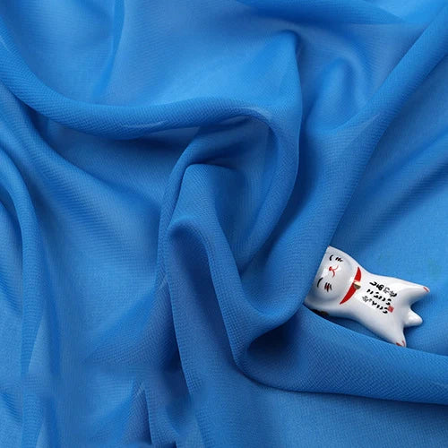 Une Mousseline légère bleue transparente au mètre de Longan Craft France est drapée en plis souples, recouvrant partiellement une petite figurine de chat en céramique blanche peinte avec des traits de visage et une écriture japonaise.