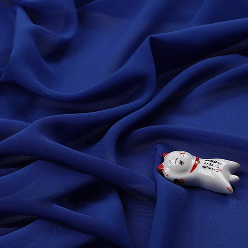 Une petite figurine de chat en céramique avec des accents rouges et noirs repose sur la "Mousseline légère transparente au mètre" de Longan Craft France, une mousseline fine et douce en tissu de polyester qui crée une toile de fond élégante et texturée.