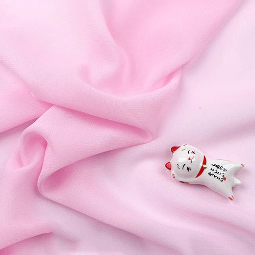 Une petite figurine de chat blanc en céramique avec des accents rouges repose sur la mousseline légère transparente au mètre de Longan Craft France, une mousseline douce et fine en tissu de polyester qui évoque le charme délicat des tenues de soirée.