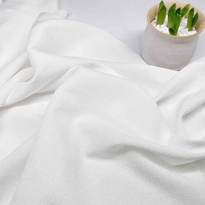 Gros plan sur le Tissu seersucker uni texturé au mètre de Longan Craft France, drapé sur une surface, avec une petite plante en pot avec des feuilles vertes et des cailloux blancs dans le coin supérieur droit.