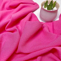 Le Tissu seersucker uni texturé au mètre de Longan Craft France, à la texture froissée et aux carreaux vichy rose vif, est exposé près d'un petit pot rose avec une succulente et des galets blancs : idéal pour des vêtements d'été frais.