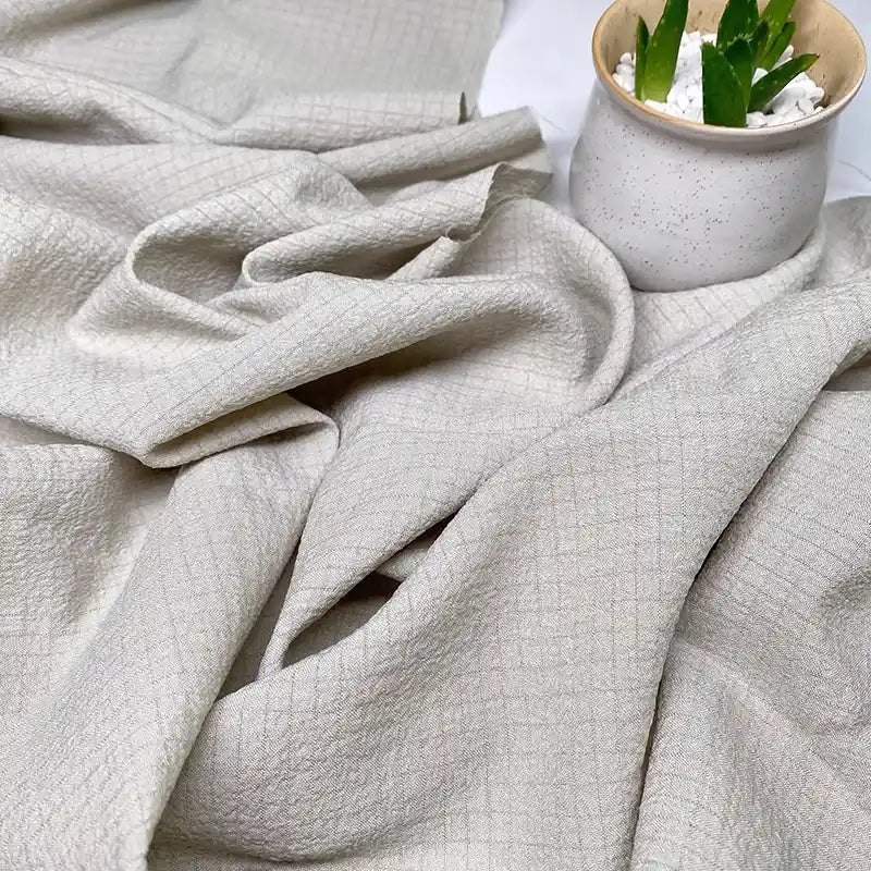 Gros plan sur le tissu polyester seersucker beige clair de Longan Craft France, drapé librement à côté d'un pot en céramique blanc moucheté contenant une petite plante succulente verte sertie de pierres blanches.
