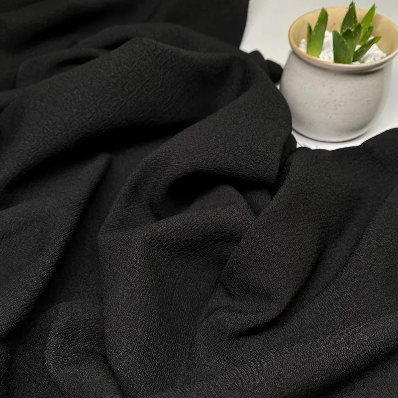 Un gros plan du Tissu seersucker uni texturé en polyester de Longan Craft France montre des plis noirs doux, avec un petit pot blanc contenant une succulente verte et des galets blancs dans le coin supérieur droit.