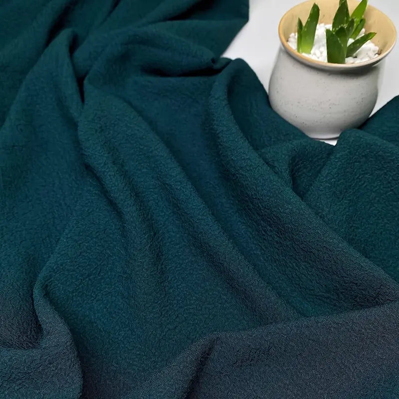 Le Tissu seersucker uni texturé au mètre de Longan Craft France en vert foncé est présenté avec des plis doux à côté d'un petit pot en céramique beige contenant une plante succulente nichée dans des pierres blanches.
