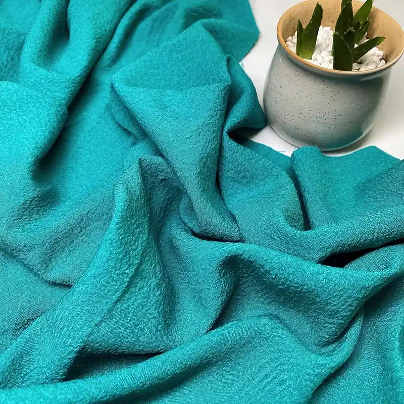 Un morceau de Tissu seersucker uni texturé au mètre de Longan Craft France est exposé à côté d'une petite jardinière grise avec une succulente verte et des galets blancs.
