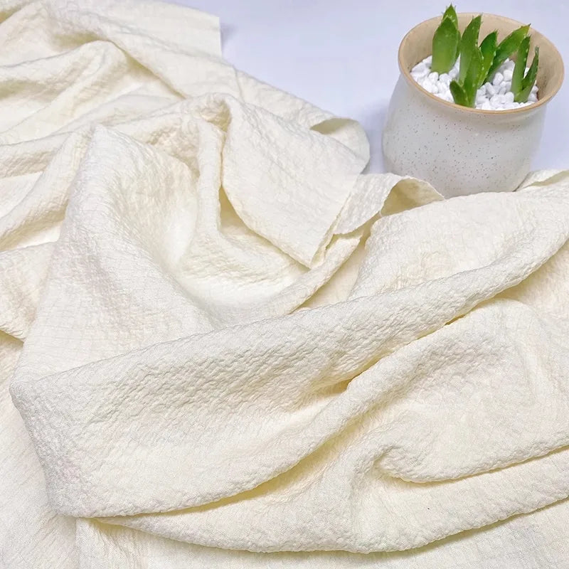 Le Tissu seersucker uni texturé au mètre de Longan Craft France, en vichy blanc cassé avec des plis texturés, est présenté à côté d'un petit pot blanc avec des plantes grasses vertes et des pierres blanches.